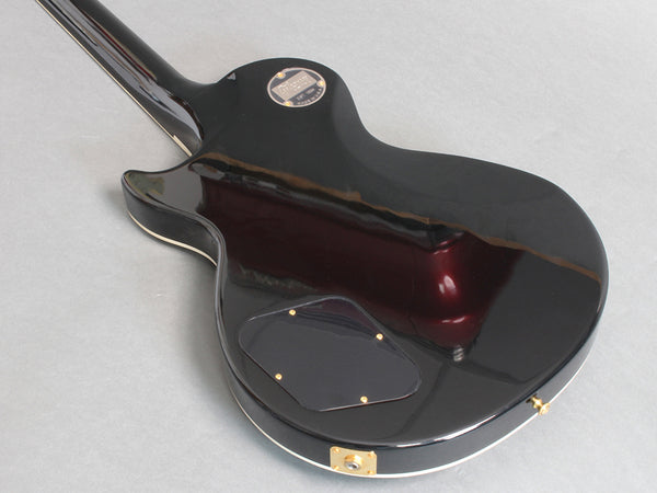 Back view of black electric guitar with glossy finish and gold hardware, showing control cavity cover and serial number plate at Coffee House Guitars