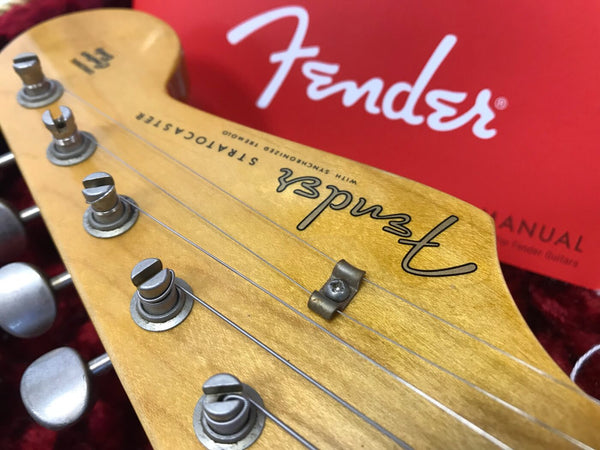 Close-up of Fender Stratocaster guitar headstock with tuning pegs and strings, featuring the Fender logo and a red Fender owner's manual in the background.