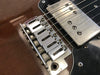 Close-up of vintage electric guitar bridge with six adjustable saddles, chrome humbucker pickup, and strings on a brown wood body with black pickguard.