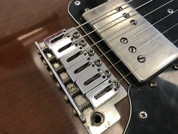 Close-up of vintage electric guitar bridge with six adjustable saddles, chrome humbucker pickup, and strings on a brown wood body with black pickguard.