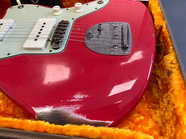 Close-up of red electric guitar body with white pickups, volume and tone knobs, and chrome bridge, showing worn paint and scratches, resting in orange plush-lined guitar case