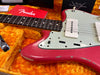 Close-up of vintage Fender electric guitar with worn pink body, white pickguard, and rosewood fretboard in orange plush-lined hard case with Fender logo accessories