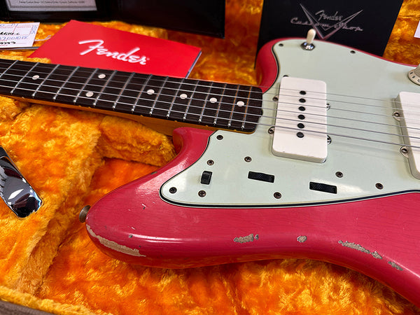Close-up of vintage Fender electric guitar with worn pink body, white pickguard, and rosewood fretboard in orange plush-lined hard case with Fender logo accessories