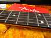 Close-up of Fender guitar fretboard with metal frets and white dot inlays on rosewood fingerboard, resting on yellow plush guitar case interior with red Fender logo tag in background.