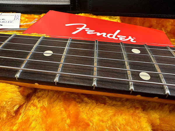 Close-up of Fender guitar fretboard with metal frets and white dot inlays on rosewood fingerboard, resting on yellow plush guitar case interior with red Fender logo tag in background.