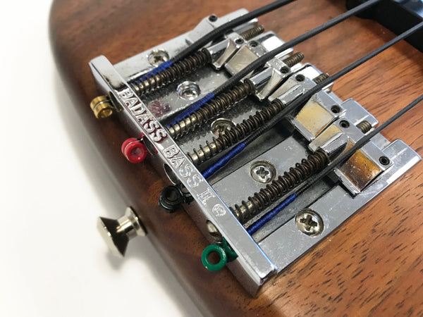 Close-up of Badass Bass II chrome bridge on wood electric bass guitar with black strings and colored string ends.