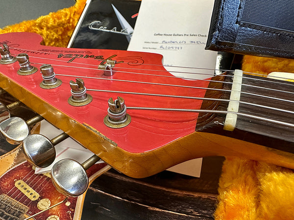 Close-up of red Fender guitar headstock with six tuning pegs, metal strings, and partial view of yellow guitar case with Certificate of Authenticity from Coffee House Guitars in the background