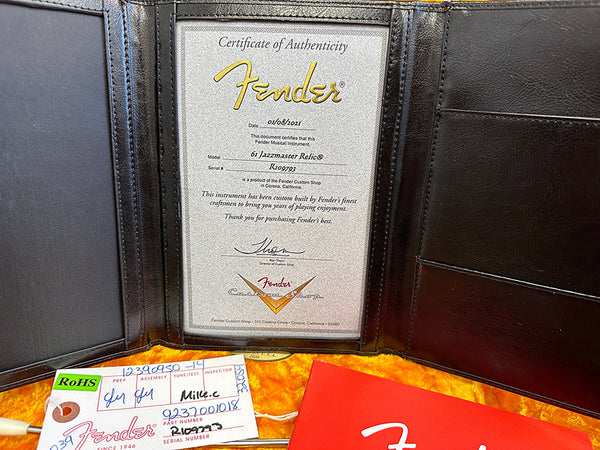 Fender Certificate of Authenticity for 61 Jazzmaster Relic guitar with serial number R100793 in black leather folder next to Fender tags and red booklet on yellow background