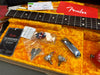 Close-up of Fender guitar neck and fretboard with accessories including keys, potentiometers, chrome tailpiece, camouflage guitar pick, and Fender instruction card inside a vintage orange plush guitar case.