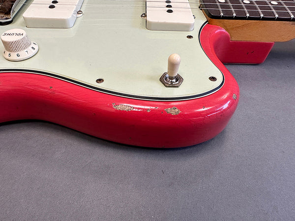Close-up of red electric guitar body with wear and paint chips, white volume knob, pickup switch, and cream pickguard on gray surface.