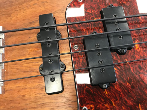 Close-up of electric bass guitar pickups and strings on a wood body with tortoiseshell pickguard, showing black pickups and black strings.