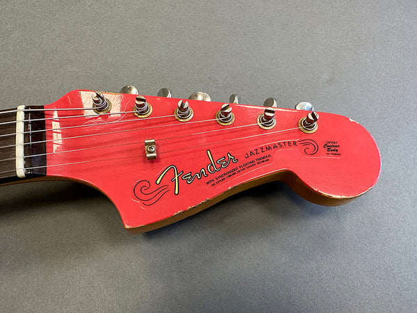 Close-up of red Fender Jazzmaster guitar headstock with six tuning pegs and visible wear on edges over gray background