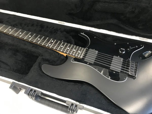 Matte black electric guitar with dual humbucker pickups, black pickguard, and volume control knob inside black plush-lined hard case, close-up of fretboard and body.