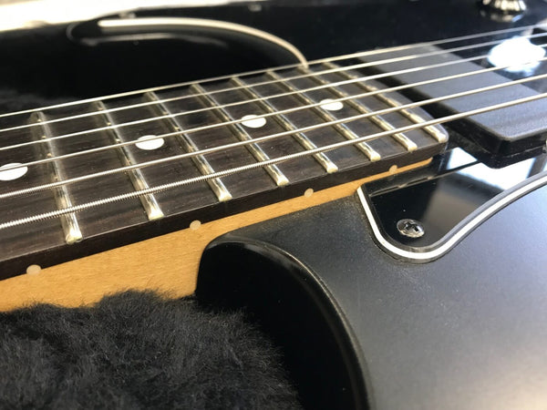 Close-up of black electric guitar fretboard with metal frets and white dot inlays on natural wood neck in soft black padded guitar case