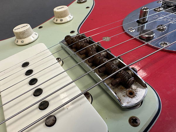 Close-up of vintage electric guitar bridge, pickups, and control knobs on red and pale green body with rusted metal parts and guitar strings.