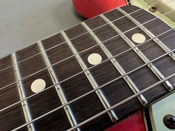 Close-up of electric guitar rosewood fretboard with metal frets and white dot inlays, showing six strings and part of red guitar body.