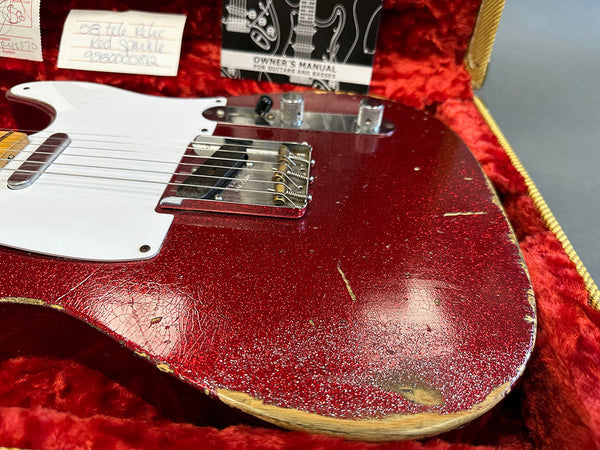 Close-up of a red 1958 Fender Telecaster electric guitar with worn and relic finish, white pickguard, chrome bridge and control knobs, resting in a red velvet-lined guitar case with owner's manual and notes in the background.