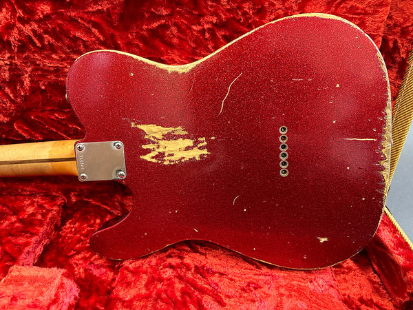 Red electric guitar body with worn and chipped paint finish, showing natural wood underneath, lying on red plush guitar case lining with visible string ferrules and neck plate screws.
