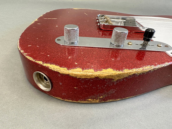 Close-up of red electric guitar body with worn and chipped finish, showing silver control knobs, tone switch, and chrome bridge on gray background, vintage relic style guitar.