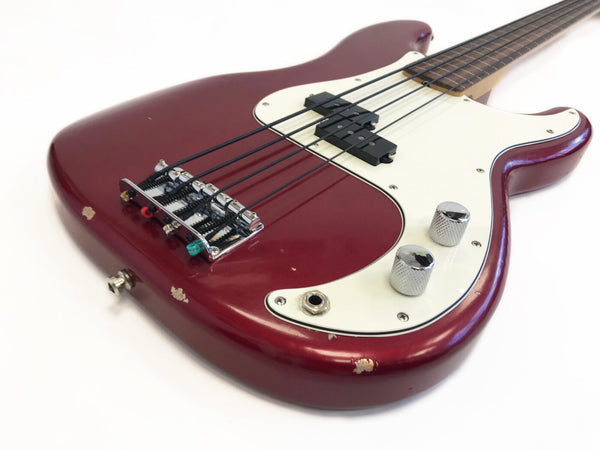 Close-up view of a red electric bass guitar with white pickguard, chrome bridge and control knobs, showing signs of wear and paint chipping on the body.