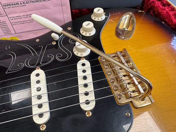 Close-up of electric guitar bridge, white pickups, tone and volume knobs, and tremolo arm on sunburst finish guitar body with partial pink hardware and electronics notes in background