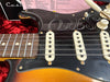 Close-up of a vintage Stratocaster-style electric guitar with relic finish, showing worn black pickguard, cream single-coil pickups, volume and tone knobs, and part of the rosewood fretboard.