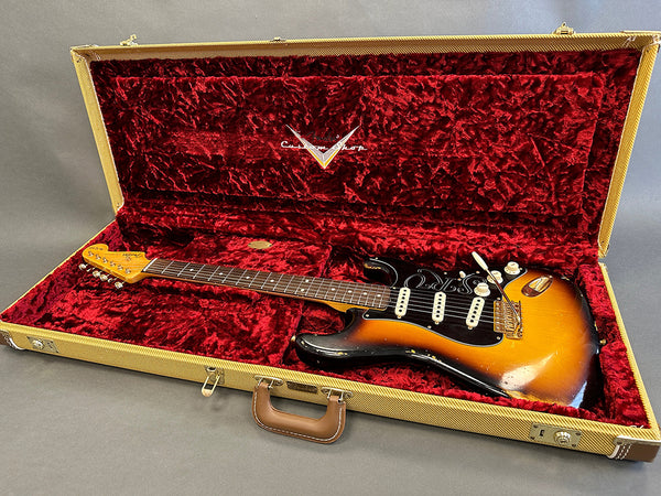Vintage Fender Stratocaster electric guitar in sunburst finish with black pickguard and signature etched design, displayed inside open tweed hard case with red plush interior lining.
