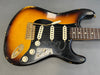 Vintage sunburst electric guitar with black pickguard featuring "SRV" engraving, three single-coil pickups, gold bridge and hardware, and white control knobs on gray background