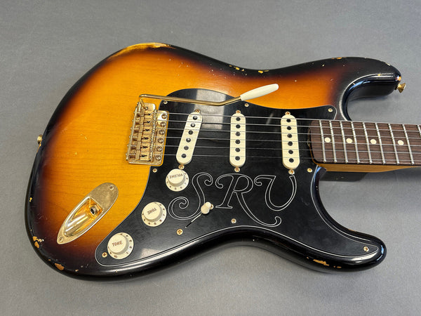 Vintage sunburst electric guitar with black pickguard featuring "SRV" initials, three single-coil pickups, and white control knobs on gray background