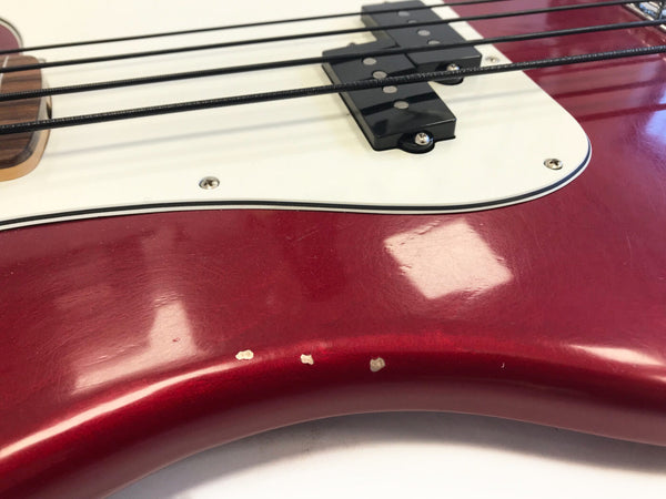 Close-up of red electric bass guitar body showing white pickguard, black pickups, and visible paint chips on the lower edge.