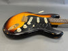 Vintage sunburst electric guitar with worn body finish and black pickguard featuring "SRV" engraving, three cream single-coil pickups, white tone and volume knobs, and tremolo arm