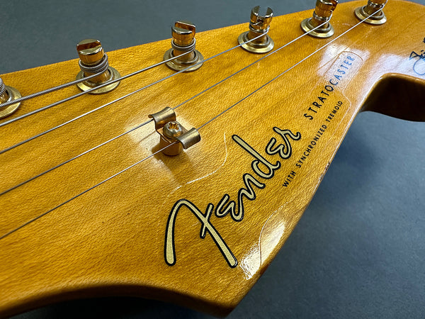 Close-up of vintage Fender Stratocaster headstock with gold tuning pegs and strings, natural wood finish, and black Fender logo on light background