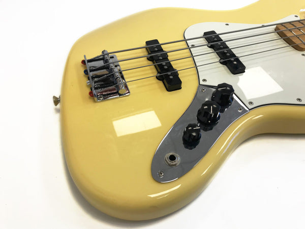 Close-up of cream-colored electric bass guitar body with chrome bridge, black pickups, white pickguard, and three black control knobs on reflective control plate
