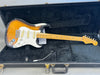 Fender Stratocaster electric guitar with sunburst finish, maple neck, and three single-coil pickups in black hard case with tremolo arm and control knobs visible
