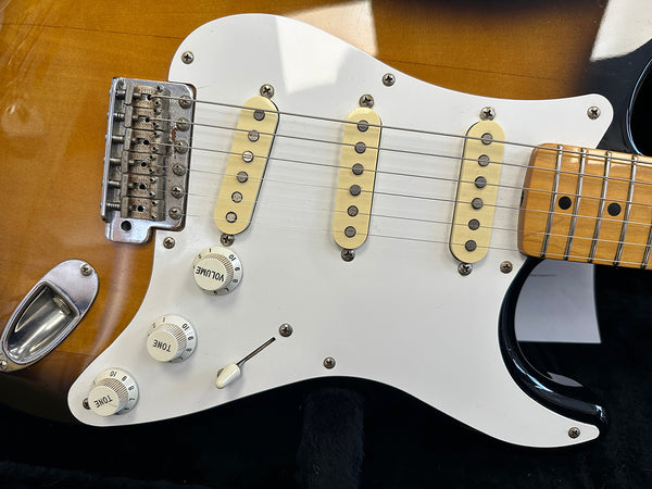 Close-up of electric guitar body with sunburst finish, white pickguard, three single-coil pickups, volume and tone control knobs, and tremolo bridge
