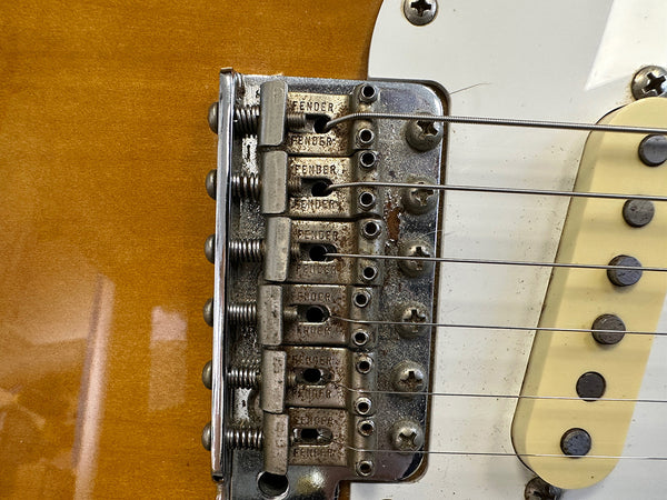 Close-up of vintage Fender guitar bridge, showing rusted metal saddles and six steel strings on honey burst finish guitar body.
