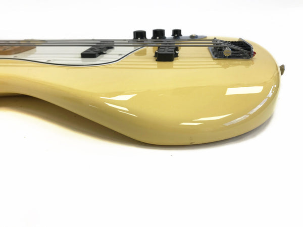 Close-up side view of cream-colored electric bass guitar body with black pickups, chrome bridge, and control knobs on white background