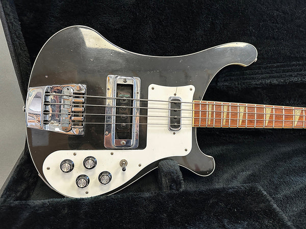 Black electric bass guitar with white pickguard and four control knobs resting in a black plush case