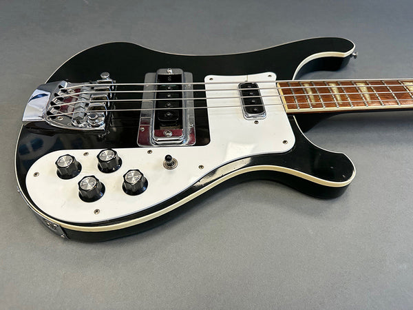 Black electric bass guitar with white pickguard and chrome hardware on gray background, close-up of control knobs and pickups