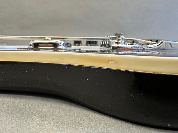 Close-up of black electric guitar body edge with cream binding and chrome bridge hardware, showing minor wear detail