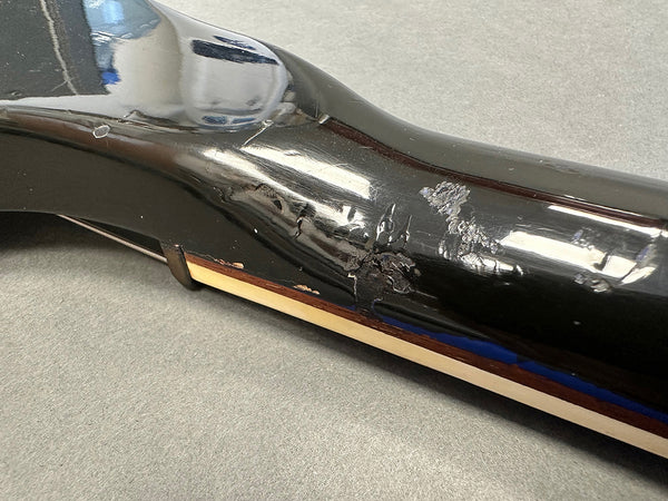 Close-up of black guitar neck heel showing wear, scratches, and chips in the finish with cream binding and rosewood strip on a gray surface