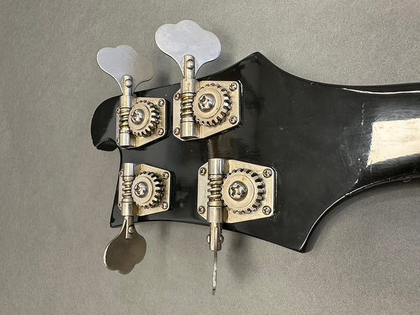 Close-up view of black bass guitar headstock showing four silver tuning pegs and gear mechanisms on gray background