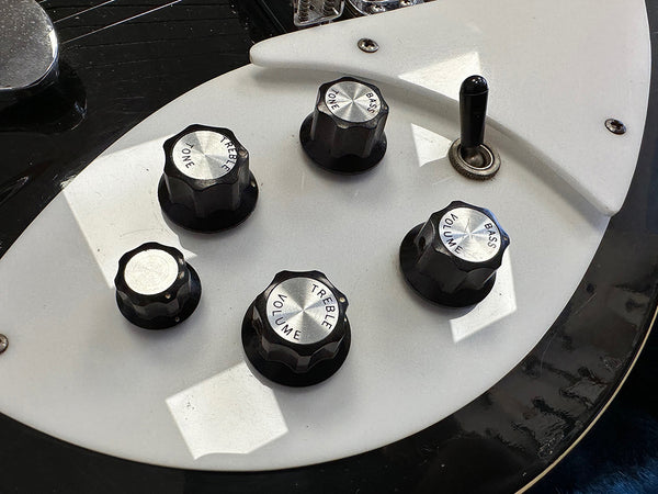 Close-up of black and silver guitar control knobs on white pickguard with toggles for volume, treble, tone, and bass on electric guitar body