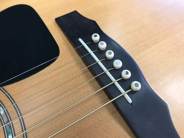 Close-up of acoustic guitar bridge with black pickguard, white bridge pins with abalone dots, and steel strings on natural wood guitar top
