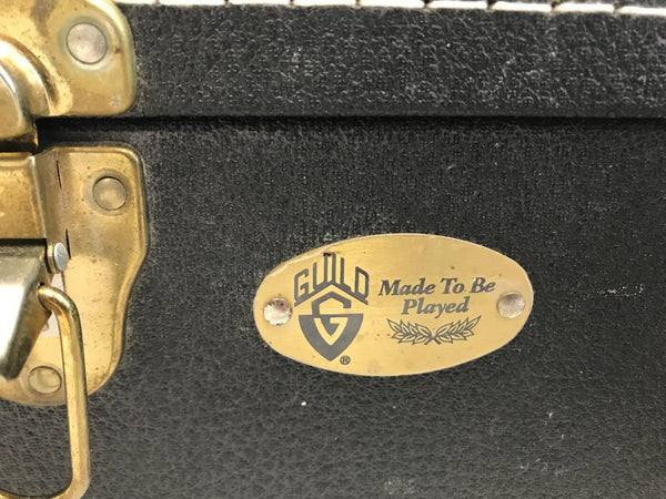 Close-up of black guitar case latch with brass Guild logo plate and "Made To Be Played" engraving on the front