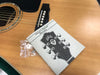 Close-up of acoustic guitar body with owner's manual and warranty card placed on top, showing guitar bridge and tuning keys illustration on manual cover, with small plastic bag containing guitar accessories.