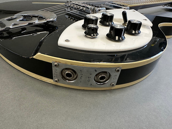 Close-up of black electric guitar body showing white pickguard, black control knobs, chrome bridge, and two metal input jacks with serial number 9621 plate on gray surface.