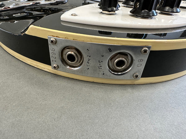Close-up of vintage electric guitar dual output jack plate with screws on black guitar body and cream binding edge.