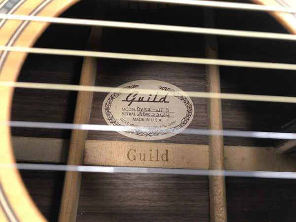 Close-up of Guild acoustic guitar interior label showing model DV52-NT S and serial number ADS22614 made in USA with visible wooden bracing and guitar strings in foreground
