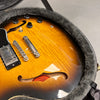 Close-up of sunburst finish semi-hollow electric guitar body with black control knobs and chrome bridge inside gray plush-lined guitar case